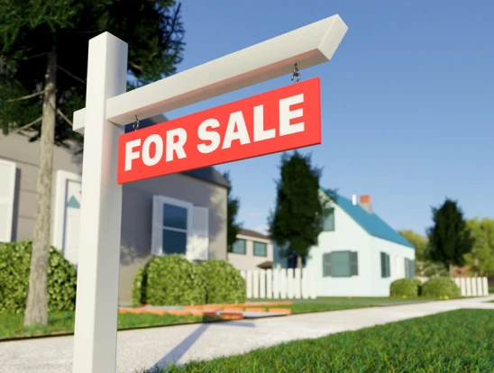 A for sale sign stands in front of a house, indicating it is available for purchase.
