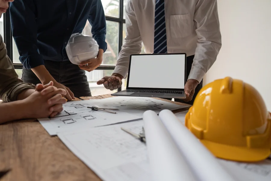 Three individuals in hard hats are focused on a laptop screen, collaborating on a project or discussing information.